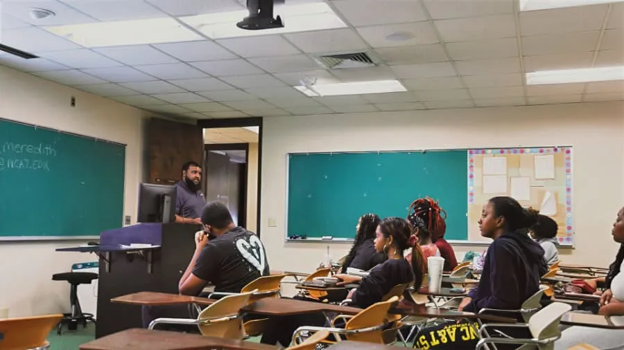 A teacher stands at a podium in front of a classroom, addressing students seated at desks. Some students have notebooks and drinks on their desks. The room has fluorescent lighting, green chalkboards, and a bulletin board.
