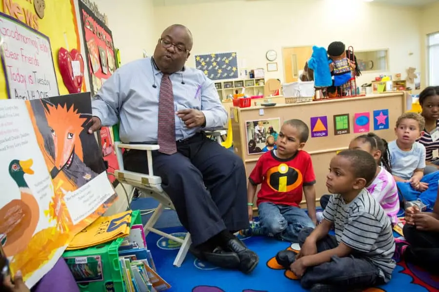 A man in glasses reads a picture book to a group of young children sitting on a colorful classroom rug, with bright decorations and educational materials in the background.