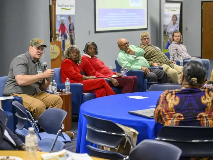 A diverse group of people sit on a panel in a conference room, engaging in a discussion. A man in a cap speaks into a microphone, while others listen. A presentation screen and banner are visible in the background. The room has blue-covered tables.