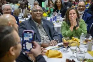 Man and woman seated at a banquet table, reacting with joy and surprise, surrounded by smiling attendees; the woman covers her mouth with her hands while the man smiles warmly, as someone in the foreground captures the moment on a phone.