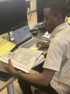A man sits at a desk using a laptop while holding and reading a large document or folder. Multiple computer monitors, papers, and a phone are on the desk. The workspace appears to be an office or computer lab.