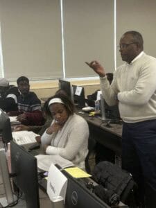 A man stands and gestures while speaking to students seated at computers in a classroom. One student appears focused on paperwork, while others look toward the instructor. Computer monitors and documents are visible on the desks.