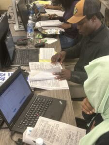 Several people sit at a long table working on laptops with papers, food containers, notebooks, and water bottles beside them. One person in the foreground examines printed documents.