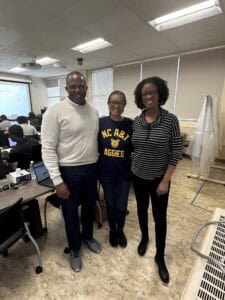 Three people stand smiling in a classroom. One person in the center wears an NC A&T Aggies shirt. Desks, students, and a projection screen are visible in the background.