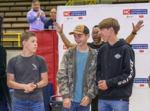 Three teenage boys stand together smiling at an indoor event, while people in the background applaud and cheer. The backdrop features “NC Cooperative Extension” logos.