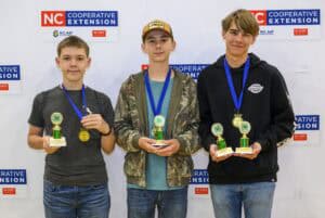 Three teenage boys standing side by side, each wearing a blue ribbon with a gold medal and holding a green and gold trophy, in front of a backdrop with NC Cooperative Extension logos.