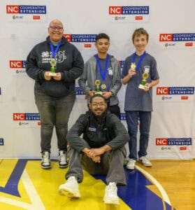 Three smiling students wearing medals and holding trophies stand in front of an NC Cooperative Extension backdrop; an adult kneels in front of them on a gym floor, also smiling at the camera.