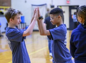 Two boys wearing safety goggles and matching blue shirts touch palms together in a gymnasium, engaging in a team-building activity. Another child stands nearby, watching.
