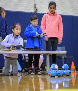 Three children interact with a small blue robot on the floor in a gymnasium. Two of the children hold tablets, likely controlling the robot, while the third watches attentively. An orange cone is nearby.