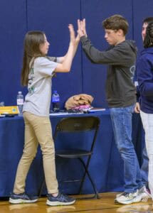 Two teens stand facing each other, touching hands as if playing a game. They are in a gym with a folded chair and table holding drinks and bags behind them. Both look focused and engaged in the activity.