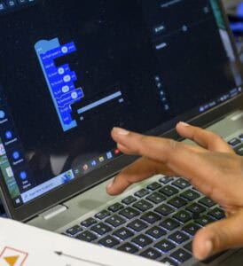 A person’s hand hovers over a laptop keyboard. The laptop screen displays a block-based coding interface with code blocks for a project, likely using a program designed to teach coding to beginners.