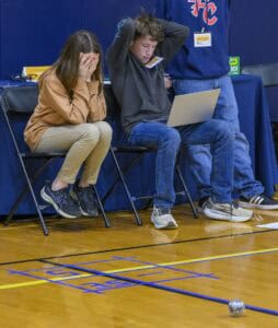 Two students sit on chairs in a gym; one covers her face in stress while the other looks frustrated at a laptop. A grid is taped on the floor with a small robot ball resting inside it.