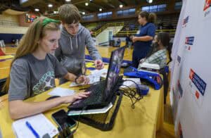 Two students work together on a laptop at a table in a gymnasium, checking papers, while others are seated or standing nearby; a competition banner is visible on the right.