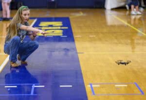 A young woman crouches on a gym floor, reaching toward a small flying drone hovering nearby. Blue tape marks and gymnasium lines are visible on the polished wood floor.