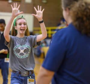 A young woman smiles excitedly with her hands raised, wearing a green headband and a The Gamecprones Union County 4-H T-shirt, facing another person in a navy shirt inside a gymnasium.