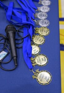 A row of gold and silver medals with blue ribbons labeled “GAME OF DROIDS 1st Place” on a blue table next to a black microphone.