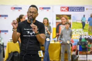 A man wearing glasses and a black T-shirt speaks into a microphone at an indoor event. People stand and work at tables in the background, with NC Cooperative Extension banners visible behind them.