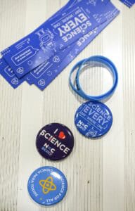 A set of blue science-themed event wristbands, blue and dark purple buttons with science messages, and a blue silicone bracelet, all arranged on a white striped surface.