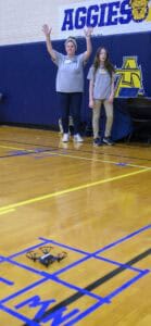 Two students stand on a gym floor near a table and banner reading AGGIES. One student raises both arms as a small drone hovers above a blue-marked starting area on the floor.