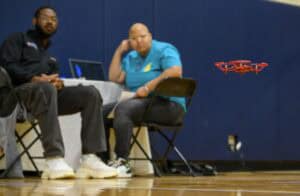 A small red drone hovers indoors near two seated people, one with a laptop on a table, both watching the drone. The setting appears to be a gymnasium with blue padded walls.
