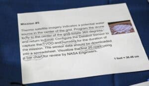 A printed instruction sheet titled Mission #5 describes a drone task, with handwritten circles around words like fly, base, TVOC, humidity, first 20 rows, and bar chart. A small photo and ruler are visible.
