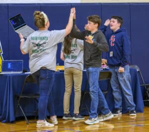 Four teens celebrate in a gym, high-fiving. One holds a laptop, and two wear Nash County Drone Team shirts. They stand by tables with tech equipment, smiling and excited.