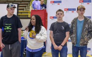 A woman speaks into a microphone while standing with three teenage boys in casual clothes at an indoor event. Behind them is an NC Cooperative Extension banner.