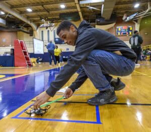 A young man crouches on a gym floor, adjusting a small drone placed inside a blue taped square, during a school event. Other people and equipment are visible in the background.
