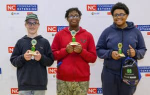 Three smiling teens stand side by side holding green 4-H trophies in front of an NC Cooperative Extension backdrop. The middle person wears a red hoodie, and the others wear dark sweatshirts.