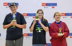Three smiling children stand in front of an NC Cooperative Extension banner, each holding a trophy or medal. Two children wear black t-shirts and one wears a red striped shirt. All have awards and blue ribbons around their necks.