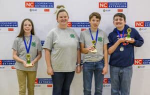 Four smiling youth stand in front of an NC Cooperative Extension backdrop. Three wear medals and hold trophies, and one wears a gray 4-H shirt. They appear to be celebrating a recent achievement or competition.