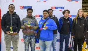 Six young people stand in front of an NC Cooperative Extension backdrop, holding trophies and smiling at the camera at an indoor event.