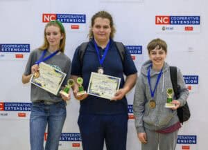 Three smiling teens stand in front of an NC Cooperative Extension banner, each holding certificates, trophies, and wearing medals around their necks, celebrating their achievements.