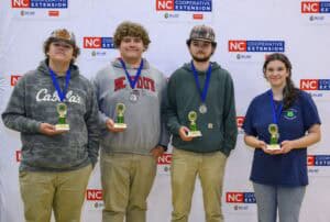 Four young people stand in front of an NC Cooperative Extension banner, each holding a trophy and wearing a medal around their neck. They are smiling and posing for a group photo.