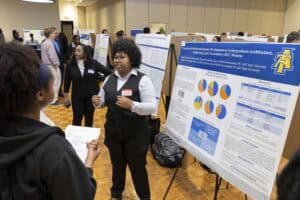 A woman in business attire explains research displayed on a poster board featuring charts and graphs to another woman at an academic conference with multiple research posters and attendees in the background.
