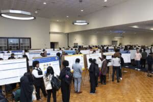 A large room filled with people viewing and discussing research posters displayed on easels; attendees are engaged in conversation and examining charts and data presentations.