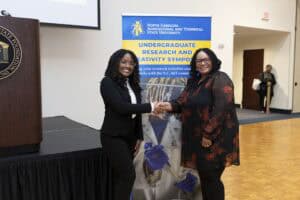 Two women smile and shake hands in front of a North Carolina A&T State University Undergraduate Research and Creativity Symposium banner in a conference room setting.