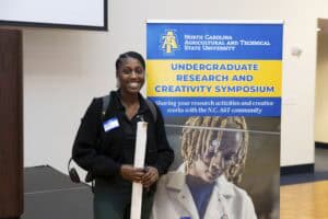 A smiling woman stands indoors holding a rolled-up poster in front of a banner for the North Carolina A&T State University Undergraduate Research and Creativity Symposium.