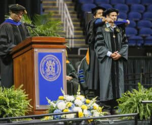 A graduate stands on stage while being hooded by faculty during a graduation ceremony. Another official speaks at a podium adorned with a university seal. Flowers and green plants decorate the stage.