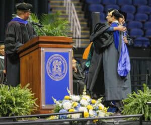 A graduate in cap and gown hugs a faculty member on stage at a graduation ceremony, while another faculty member stands at a podium. Blue and yellow flowers and university seal are visible in the foreground.