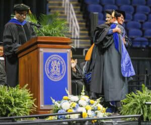 A graduation ceremony: a graduate in cap and gown hugs a faculty member on stage while another official stands at a podium. The stage is decorated with flowers and ferns, and empty blue seats are visible in the background.