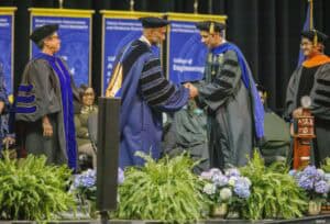 A graduate in academic regalia shakes hands with a faculty member on stage at a commencement ceremony, with other faculty and banners in the background and green plants in the foreground.