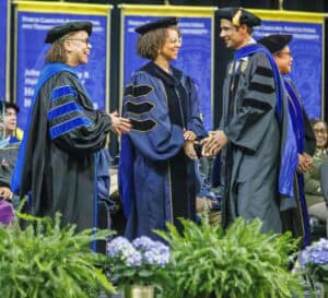 Four people in academic regalia stand onstage at a graduation ceremony, smiling and shaking hands. Blue banners and an arrangement of green plants and purple flowers are visible in the foreground.