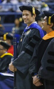 A smiling graduate in a cap and gown holds a diploma case while standing next to another person in academic regalia at a graduation ceremony.