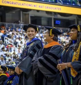 Three graduates in academic regalia stand and smile during a commencement ceremony. A large screen in the background reads CONGRATULATIONS NC A&T GRADUATES, and the audience is seated behind them.