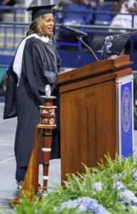 A woman in a graduation cap and gown speaks at a podium during a commencement ceremony. A ceremonial mace stands beside the podium, and flowers decorate the stage.
