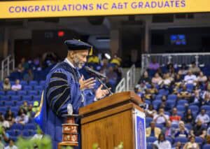 A speaker in academic regalia addresses graduates from a podium at an NC A&T commencement ceremony, with Congratulations NC A&T Graduates displayed on a large banner in the background.