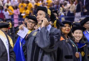 A man in academic regalia talks on a cellphone at a graduation ceremony, surrounded by other faculty members in caps and gowns, with a crowd of people in the background.