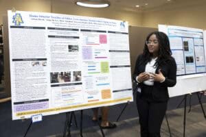 A woman stands beside a large academic research poster titled Home Interior Textiles of Older, Low-income Homeowners Aging in Place. She presents her work at a conference, with another research poster visible in the background.