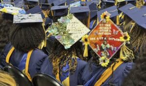 Graduates in caps and gowns sit in rows; some mortarboards are decorated with flowers, quotes, and colorful designs, celebrating their achievements at a graduation ceremony.